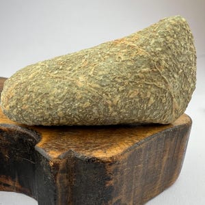 May include: A smooth, light green and brown stone with a rough texture. The stone is resting on a wooden base.