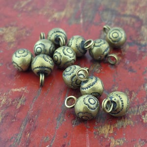 May include: A collection of small, spherical brass charms, each featuring a central loop for attachment. The surface of every charm is decorated with a repeating, etched floral pattern consisting of dark, recessed lines against the metallic gold-toned brass.