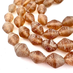 May include: A close-up of a strand of brown, barrel-shaped beads. The beads have a frosted, textured appearance with white speckling. The beads are arranged on a white background. The beads are a warm, earthy tone.