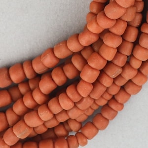 May include: A strand of terracotta-coloured glass beads. The beads are small and round, with a slightly irregular shape. They are strung together on a clear thread.