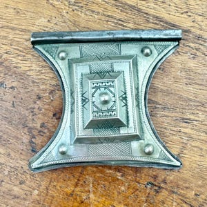 May include: A silver-toned metal decorative piece with an intricate geometric design. The central square is surrounded by a series of smaller squares and detailed patterns. The piece has a curved, hourglass-like shape with rounded edges and small, circular accents.
