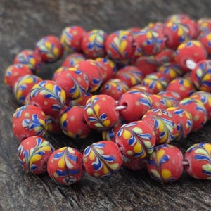 May include: A strand of red glass beads with a floral design in yellow, blue, and white. The beads are round with a hole through the center. The beads are strung together on a white string. The beads are a decorative craft supply.