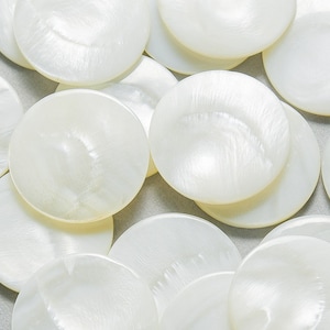 May include: A collection of round, white, mother-of-pearl buttons. The buttons have a lustrous, iridescent sheen and a smooth, slightly domed surface. The buttons are scattered across a light gray surface.