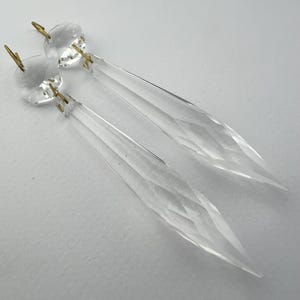 May include: Two clear crystal chandelier pendants with faceted teardrop shapes. Each pendant has a round, faceted top with a gold-colored metal pin for hanging. The pendants are displayed on a white background.