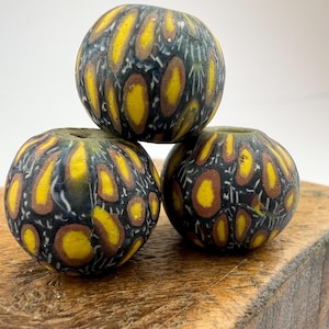 Peut inclure: Trois perles rondes en verre, avec un fond sombre et un motif de formes ovales jaunes et marron. Les perles sont empilées, avec un trou visible en haut de chacune. Artisanat.