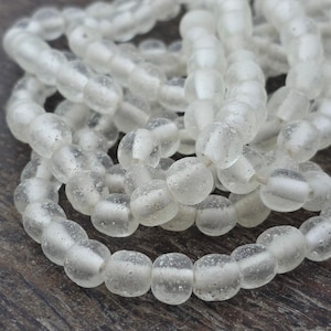 May include: A collection of translucent white glass beads with a frosted, matte texture. The beads are spherical and strung together in multiple overlapping loops, showcasing a consistent size and a slightly irregular, organic surface finish.