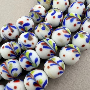 Puede incluir: Una colección de canicas de vidrio blanco con remolinos azules, rojos y amarillos. Las canicas están dispuestas en semicírculo sobre una superficie marrón claro.