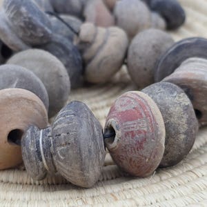 May include: A collection of weathered, stone-like beads in shades of gray, brown, and red. The beads have various shapes, including rounded and cylindrical forms, strung together. The beads are resting on a woven, beige surface.
