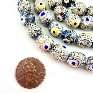 May include: Close-up of a strand of glass beads with an eye design. The beads are primarily white with a speckled pattern, and feature a blue and yellow eye in the center. A U.S. penny is included for size reference.