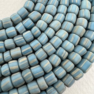 May include: A close-up of a strand of light blue glass beads with white stripes. The beads are cylindrical and have a slightly irregular shape.
