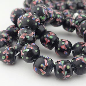 May include: Close-up of a strand of black glass beads. Each bead is decorated with a colourful floral pattern in red, green, and blue. The beads are round with a slightly flattened shape.