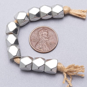 May include: A strand of silver-toned metal beads with a faceted hexagonal shape. The beads are strung on a natural-colored cord with tassels at each end. A US penny is included for scale.