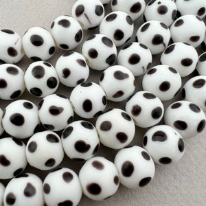 May include: A collection of white glass beads with black polka dots. The beads are round and smooth, and they are arranged in a random pattern.
