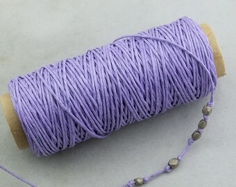 May include: A spool of lavender-colored twine with a small strand of the same color extending from the roll. A thin cord with silver-colored beads is attached to the twine. The twine is wrapped around a cardboard tube.