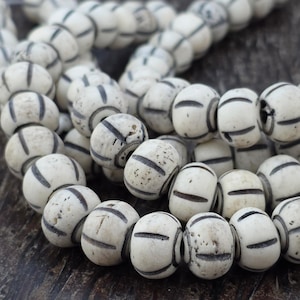 May include: A strand of round, off-white beads featuring dark, etched vertical lines that wrap around the circumference of each bead. The beads have a matte, textured surface with visible natural variations and small dark specks throughout.