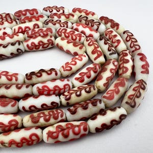 May include: A collection of white, oval-shaped beads with a red and brown leaf-like pattern. The beads are strung together, creating a necklace or bracelet. The design is symmetrical and detailed.