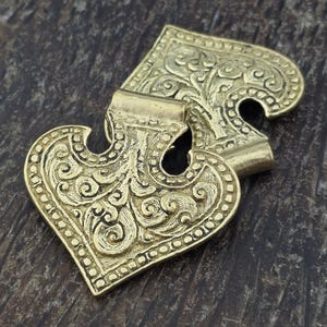 May include: A gold-toned metal cloak clasp consisting of two interlocking heart-shaped pieces. Each piece features an intricate embossed scrollwork pattern with a beaded border and a central loop for attachment, creating a decorative fastener.