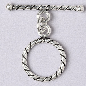 May include: A silver-toned toggle clasp with a twisted rope design. The clasp features a circular ring and a bar, connected by a series of loops. The metal has an aged, antique appearance.
