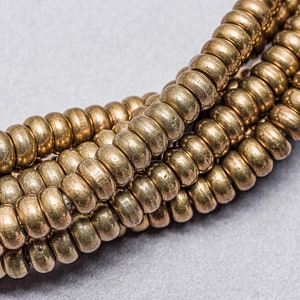 May include: A close-up of a gold-coloured beaded necklace. The beads are round and have a slightly textured surface. The necklace is layered and twisted, creating a decorative pattern.