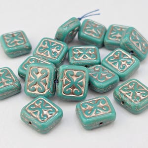 May include: A collection of square-shaped beads in a vibrant turquoise color. Each bead features a raised, metallic copper-toned embossed pattern in the center, resembling a stylized cross or knot design, with a hole for stringing.