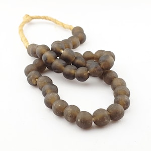 May include: A strand of dark brown, round beads with a natural, textured appearance. The beads are strung on a light brown, twisted cord, creating a necklace or decorative element. The beads have a matte finish and vary slightly in size.