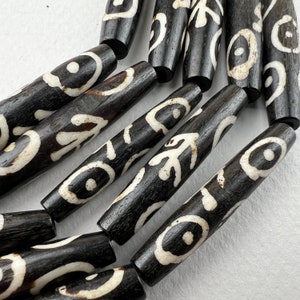 Puede incluir: Primer plano de una colecci&oacute;n de cuentas cil&iacute;ndricas de color marr&oacute;n oscuro con patrones abstractos blancos. Las cuentas est&aacute;n dispuestas en una l&iacute;nea curva, mostrando sus dise&ntilde;os y texturas &uacute;nicos. Ideal para la creaci&oacute;n de joyas.