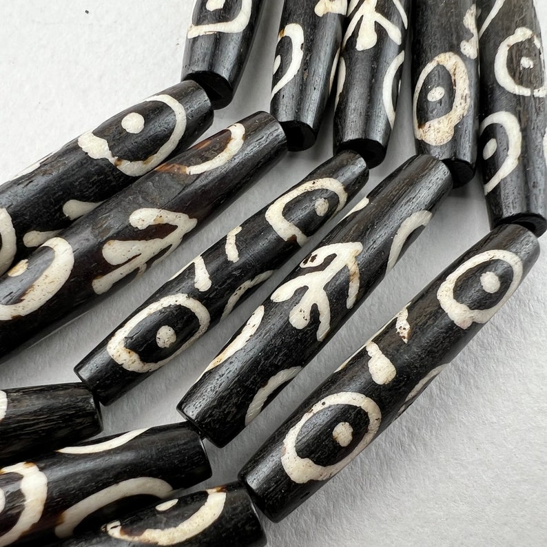 Puede incluir: Primer plano de una colecci&oacute;n de cuentas cil&iacute;ndricas de color marr&oacute;n oscuro con patrones abstractos blancos. Las cuentas est&aacute;n dispuestas en una l&iacute;nea curva, mostrando sus dise&ntilde;os y texturas &uacute;nicos. Ideal para la creaci&oacute;n de joyas.