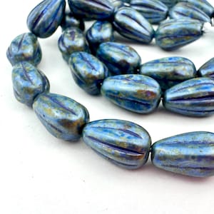May include: A strand of blue and gold-toned glass beads. Each bead is oval-shaped with vertical grooves and a metallic finish. The beads are strung together on a white string, perfect for jewelry making.