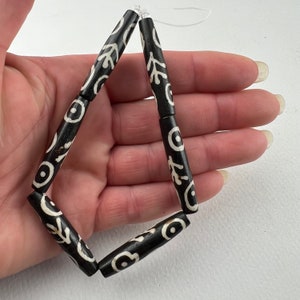 Puede incluir: Cuentas con dise&ntilde;os en blanco y negro ensartadas. Las cuentas son cil&iacute;ndricas con un fondo oscuro y dise&ntilde;os blancos estilizados. Las cuentas est&aacute;n ensartadas en un hilo transparente, formando un rombo. Las cuentas miden aproximadamente 2,5 cm de largo.