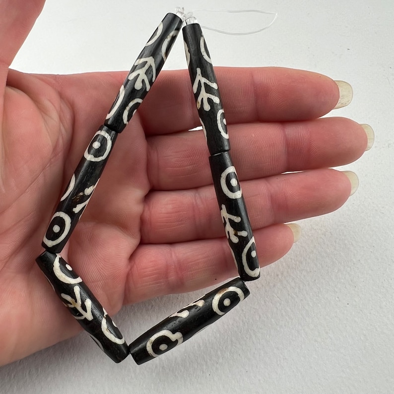 Puede incluir: Cuentas con dise&ntilde;os en blanco y negro ensartadas. Las cuentas son cil&iacute;ndricas con un fondo oscuro y dise&ntilde;os blancos estilizados. Las cuentas est&aacute;n ensartadas en un hilo transparente, formando un rombo. Las cuentas miden aproximadamente 2,5 cm de largo.
