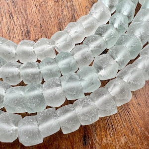 May include: A strand of frosted glass beads in a light, translucent blue color. The beads are faceted and have a slightly irregular shape, giving them a rustic and handmade look.