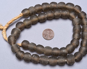 Pode incluir: Um fio de contas de vidro cinza escuro, enfiadas em um cordão de fibra natural. As contas são redondas e ligeiramente irregulares, com um acabamento fosco. Uma moeda de um centavo de dólar americano é incluída para escala.