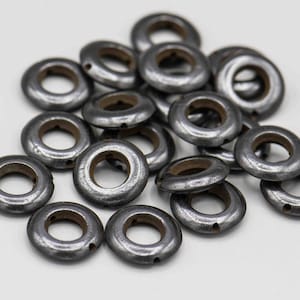 May include: A collection of small, metallic gray eyelets with a circular design and a central hole. The eyelets have a slightly weathered appearance, suggesting they are vintage or antique. They are scattered on a white surface.