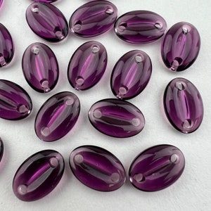 May include: A group of 15 oval-shaped, purple glass beads with two holes each. The beads are translucent and have a glossy finish.