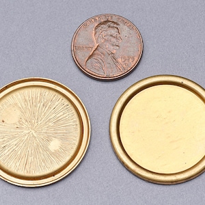 May include: Two gold-colored round metal lids with a raised edge. One lid has a textured surface with a starburst pattern. The other lid has a smooth surface. A copper penny is in the background.