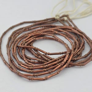 May include: A strand of metallic copper-colored tube beads with a segmented design, featuring small raised rings along the length of each bead. The beads are strung together on a thin, off-white cord, arranged in a circular, coiled pattern.