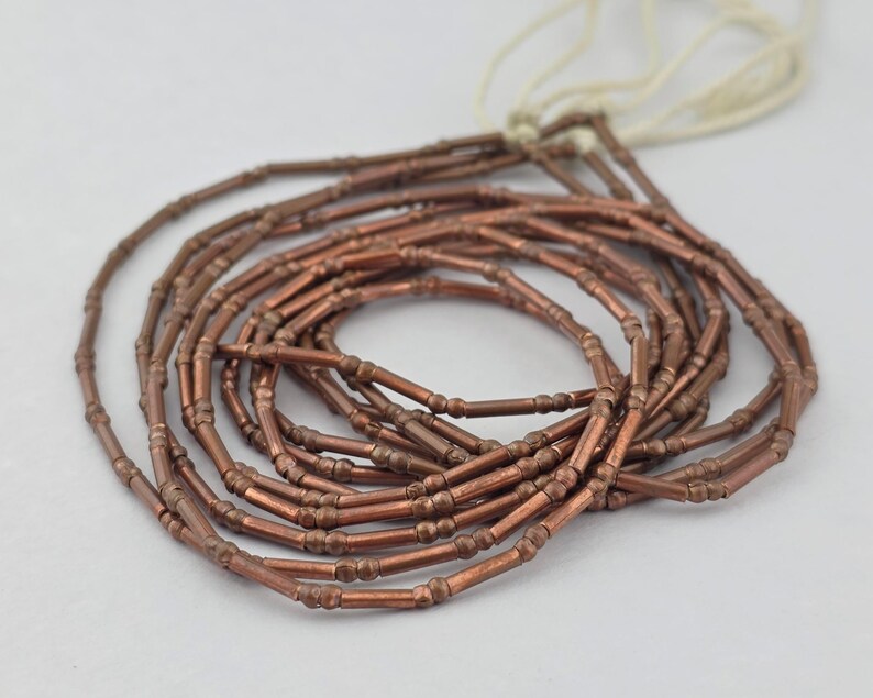 May include: A strand of metallic copper-colored tube beads with a segmented design, featuring small raised rings along the length of each bead. The beads are strung together on a thin, off-white cord, arranged in a circular, coiled pattern.