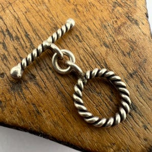 May include: Silver toggle clasp with a twisted rope design. The clasp has a round ring and a bar with rounded ends.