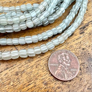 May include: Close-up of strands of light blue and white striped glass beads. The beads are rectangular with rounded edges. A U.S. penny is in the image for size reference.
