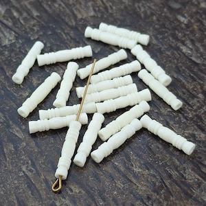 May include: A collection of off-white cylindrical beads with ribbed detailing, featuring a central spindle-like shape. One bead is threaded onto a thin, gold-toned metal wire pin with a small loop at the end, while others are scattered.
