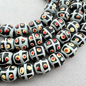 May include: A strand of oval-shaped beads with a black and white geometric pattern and red accents. The beads are made of ceramic and have a glossy finish.
