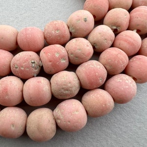 May include: A strand of pink, round, and textured beads. The beads are made of a natural material and have a rough, uneven surface.
