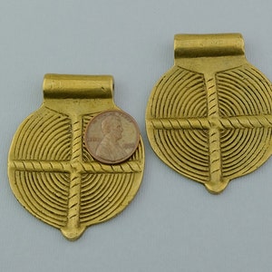 May include: Two gold-colored metal pendants with a circular design featuring concentric circles and a cross in the center. The pendants have a loop at the top for hanging.