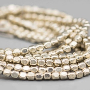 May include: A strand of silver-toned beads, each bead is faceted and has a slightly irregular shape. The beads are arranged in a loose pile, creating a textured and organic look.