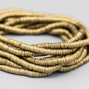 May include: Close-up of a coiled strand of golden-colored, small, round beads. The beads are tightly strung together, forming a rope-like structure. The beads have a metallic sheen, reflecting light. The background is a soft, neutral gray.