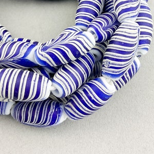 May include: Close-up of a strand of blue and white striped beads. The beads are oval-shaped with a spiral design. The beads are strung together on a white string. The background is a light gray color.