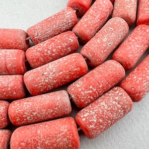 Puede incluir: Primer plano de una hebra de cuentas cilíndricas de color rojo y blanco moteado. Las cuentas están hechas de un material rugoso y texturizado y tienen un aspecto rústico y hecho a mano.