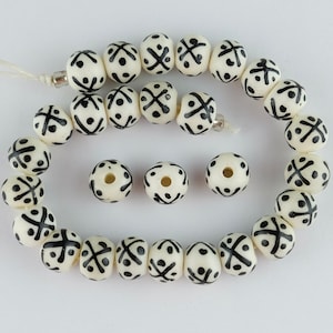 May include: A strand of white beads with black geometric patterns. The beads are strung on a white cord and there are a few extra beads to the side.