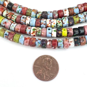 May include: Strands of colorful, patterned beads in shades of blue, red, green, and pink. The beads are cylindrical with a speckled design. A penny is included for size reference.