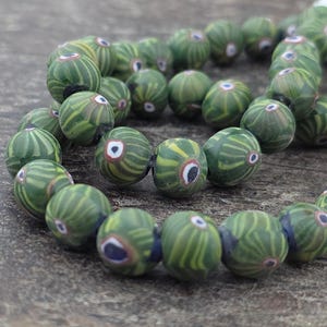 May include: A strand of green and yellow glass beads with a striped pattern. Each bead has a white and blue eye-like design. The beads are strung together on a dark string, creating a necklace or bracelet.